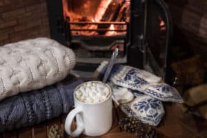 Stack of cozy knitted sweaters, handmade mittens and cup of coffee with marsh mallows on old wooden table, near burning